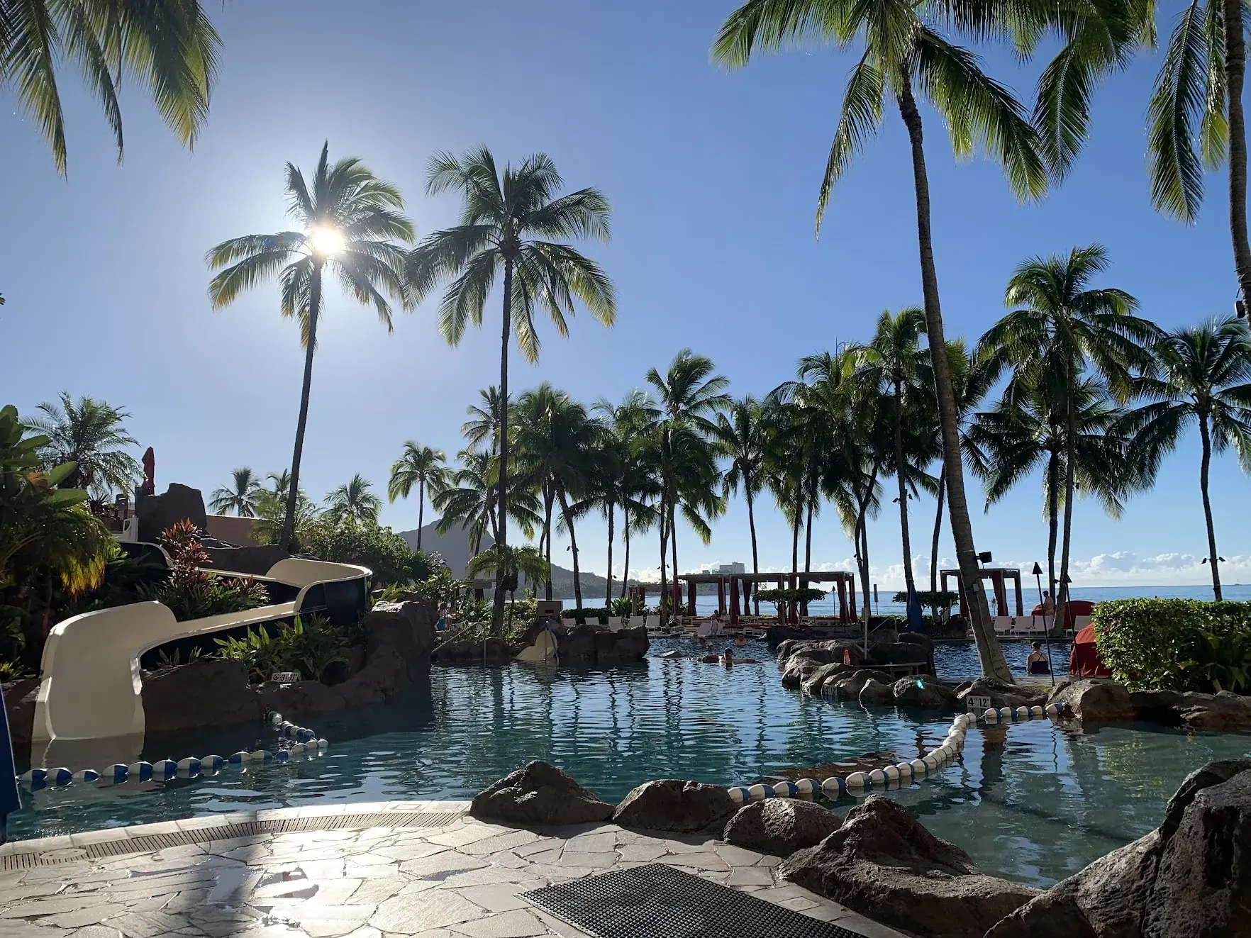 Palm trees around a resort pool with a waterslide in Waikiki, with Diamond Head in the background